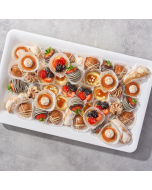 Overhead view of miniature pastries arranged in rows on a table cloth, including raspberry tulip cups, lemon cream baskets, miniature seasonal fruit tarts, miniature chocolate éclairs, and Passion fruit glazed cream puffs
