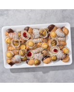 Overhead view of miniature cannoli, miniature éclairs, pistachio glazed cream puffs, miniature sfogliatelle, ricotta cheesecake tart, pignoli cookies, chocolate-dipped Amaretti cookies, arranged in rows on a stone platter