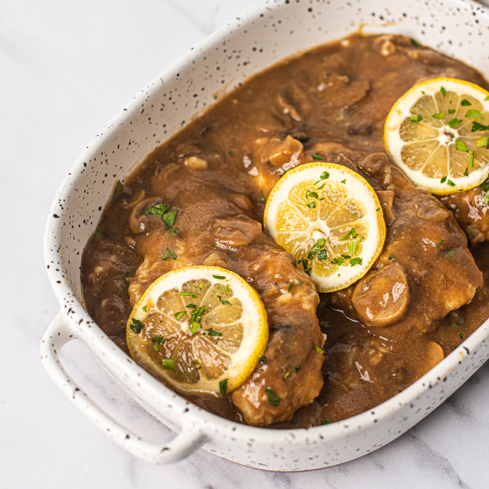 Oblique overhead view of chicken marsala, topped with rounds of lemon, served in a white stoneware dish