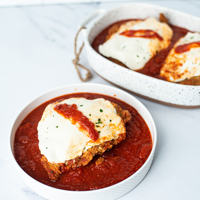 Oblique view of chicken parmigiano served in a white bowl, with a casserole dish of chicken parmigiano in the background