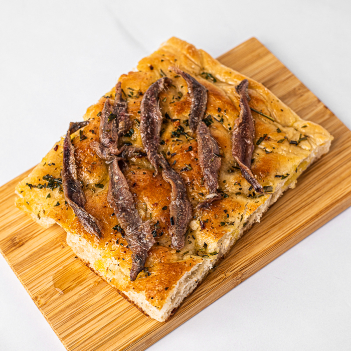 Overhead view of a corner piece of anchovy and herb-topped focaccia bread, served on a bamboo platter